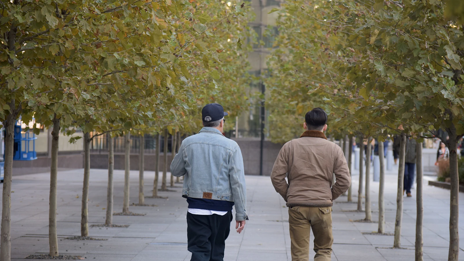 Twee mannen lopen naast elkaar over een stoep tussen de bomen.