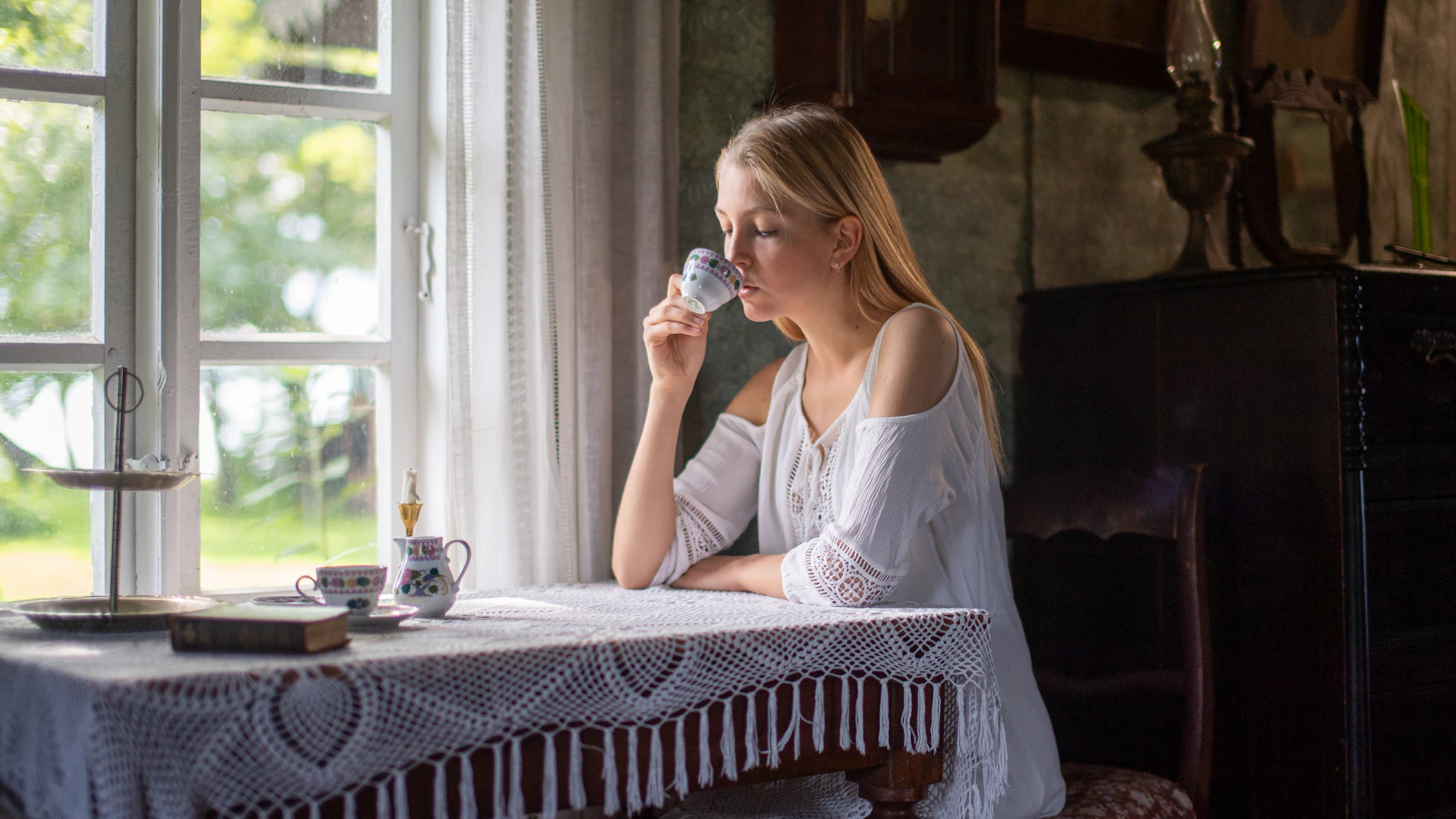 Vrouw zit met een kop thee bij het raam en kijkt rustig naar buiten.