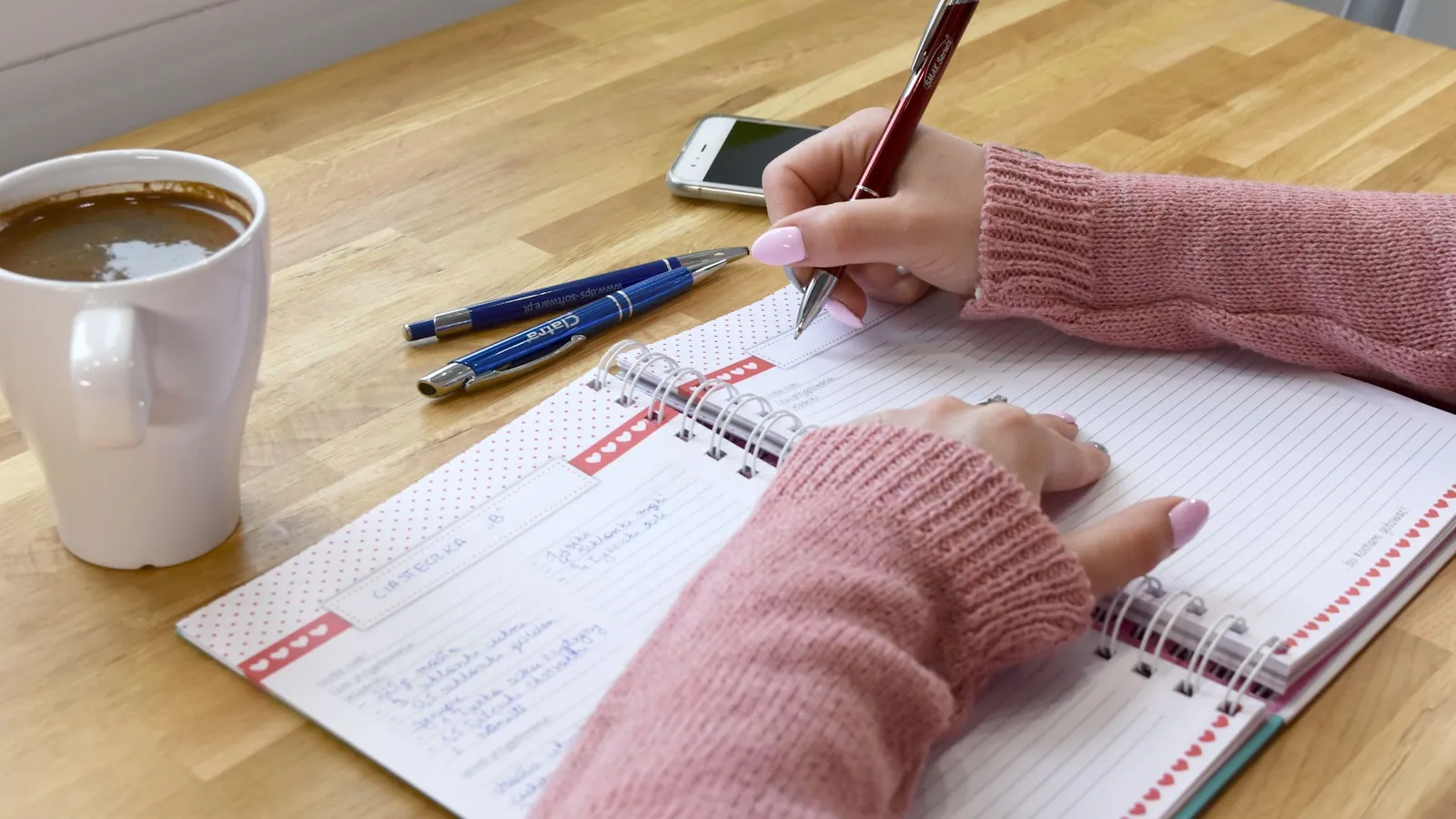 Handen schrijven in een agenda naast een kop koffie op een houten tafel.