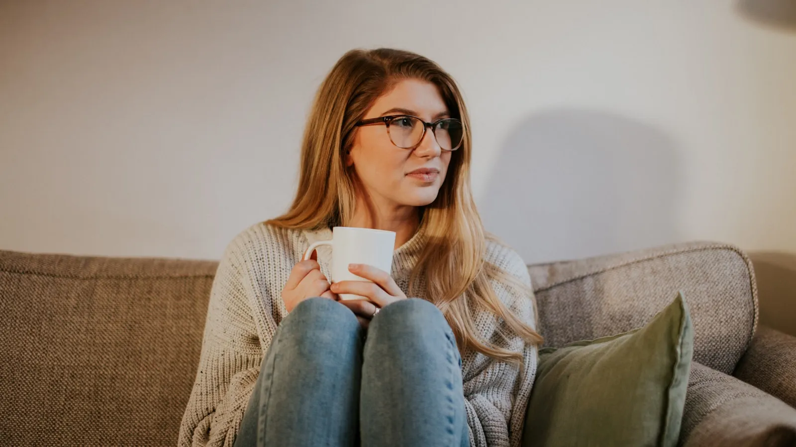 Vrouw met bril zit met een mok op de bank en kijkt rustig opzij.