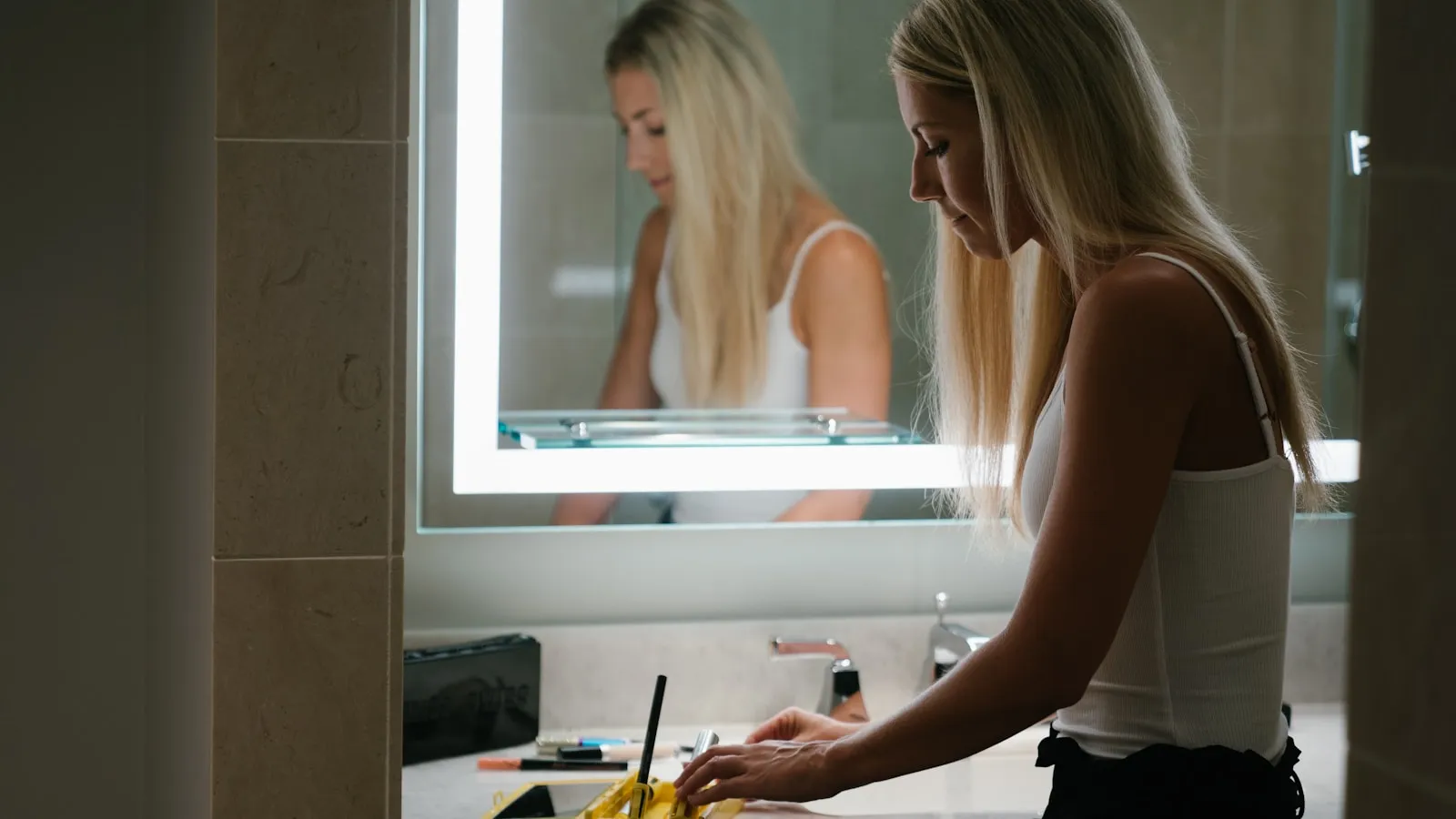 Vrouw in een wit hemdje staat bij de wastafel voor de badkamerspiegel.