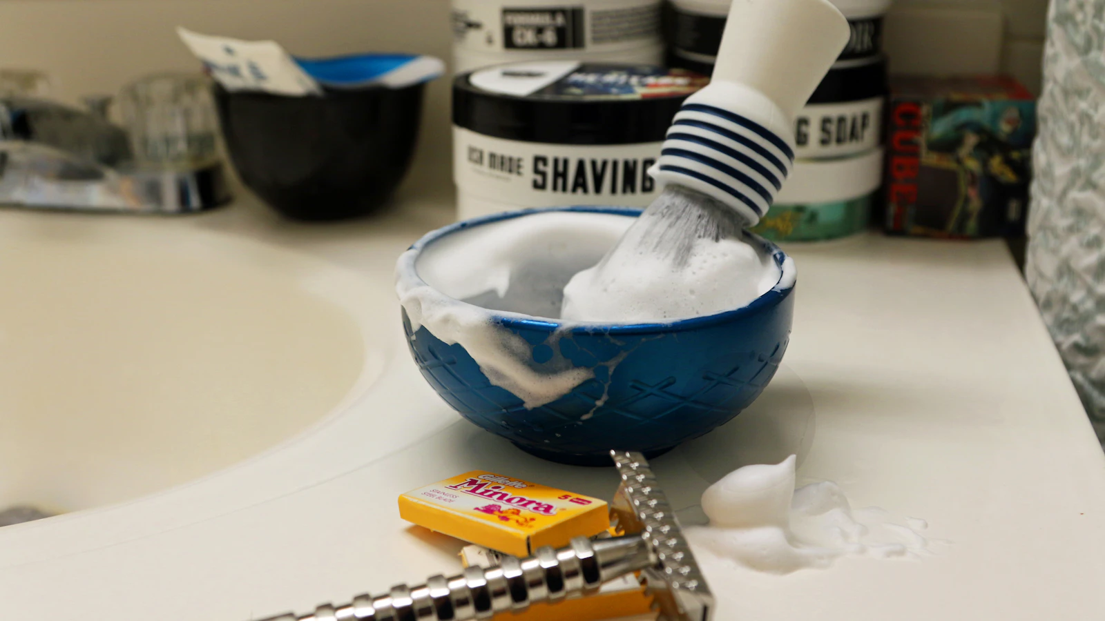 Shaving brush with lather in a blue bowl next to a safety razor on a bathroom counter.
