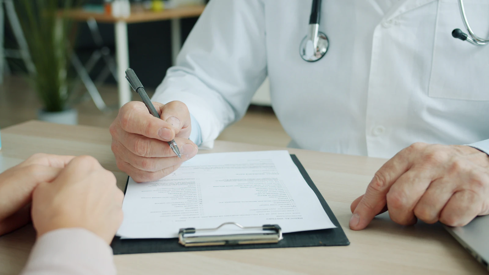 Arts vult aan een bureau een formulier in terwijl de handen van een patient tegenover hem liggen.