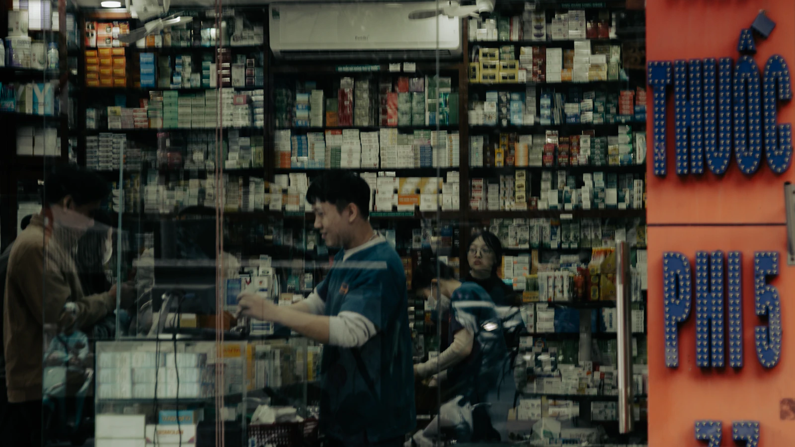 People inside a well-stocked pharmacy behind glass doors.